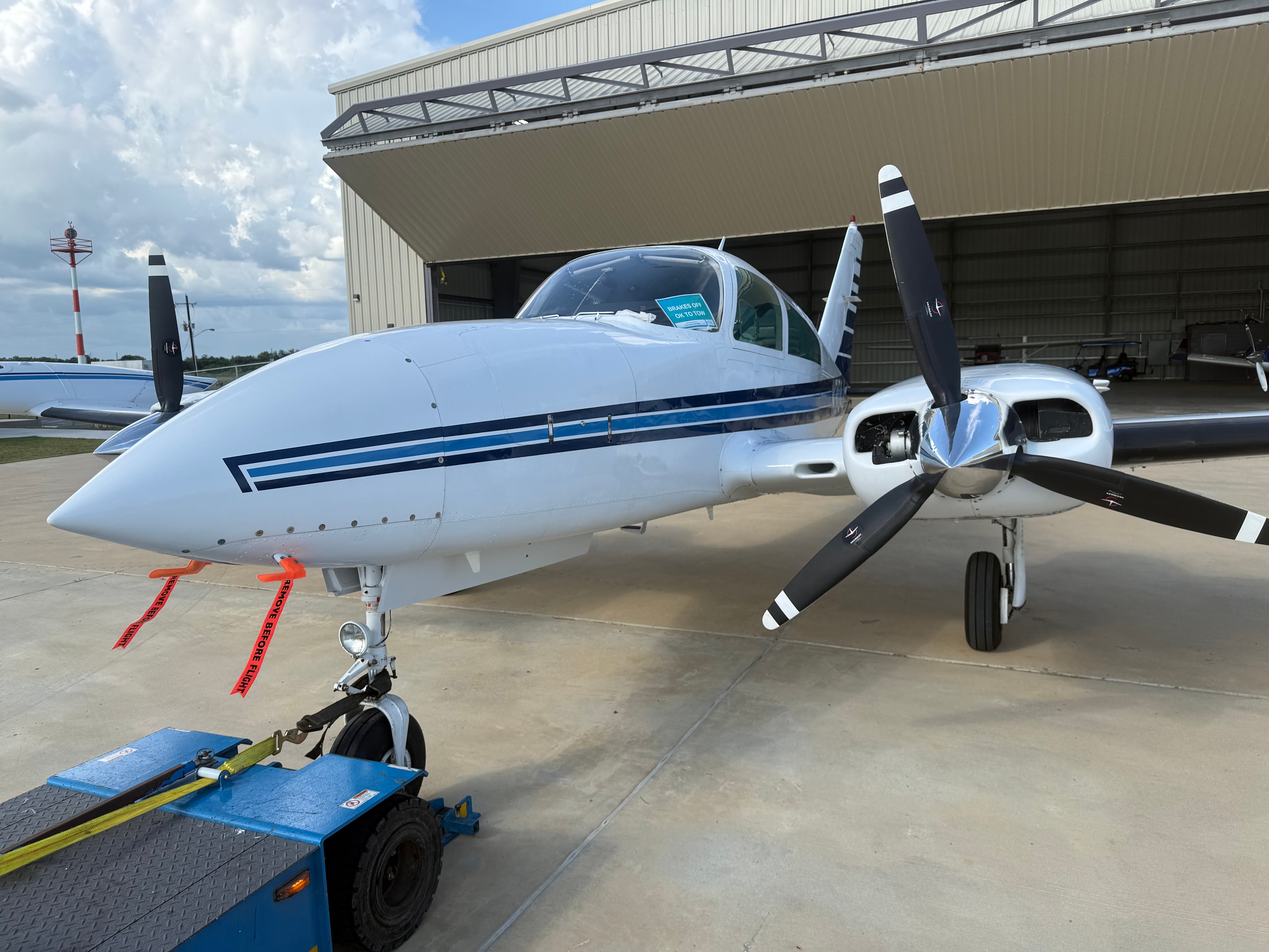 Twin-engine aircraft with polished propellers on tarmac outside hangar
